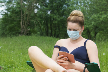 A woman is sitting in a camping chair. There is a medical mask on the face. Camping during a pandemic.