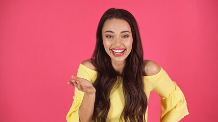 young adult woman in yellow blouse laughing and gesturing with hand isolated on pink