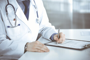 Unknown male doctor sitting and working with clipboard of medication history record in clinic at his working place, close-up. Young physician at work. Perfect medical service, medicine concept