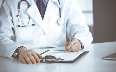 Unknown male doctor sitting and working with clipboard in clinic at his working place, close-up. Young physician at work. Perfect medical service, medicine concept