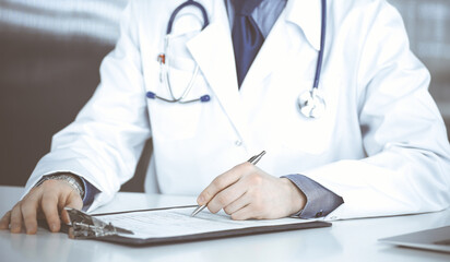 Unknown male doctor sitting and working with clipboard in clinic at his working place, close-up. Young physician at work. Perfect medical service, medicine concept