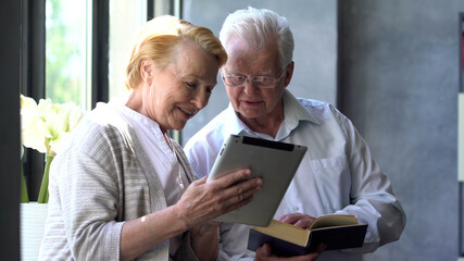 Happy lovely retired couple standing near window using digital tablet