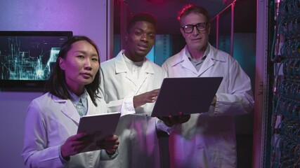 Tracking portrait shot of three IT specialists in lab coats using tablet and laptop and discussing work in data center, then looking at camera - Powered by Adobe