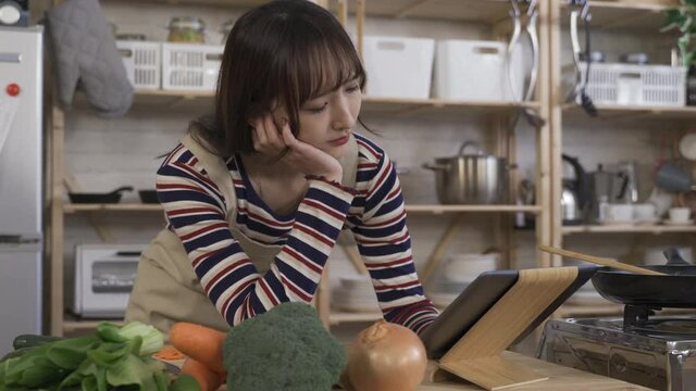 reading the steps before cooking. taiwanese young lady stooping over the tablet computer, holding head with hand by kitchen table. technology and genuine lifestyle