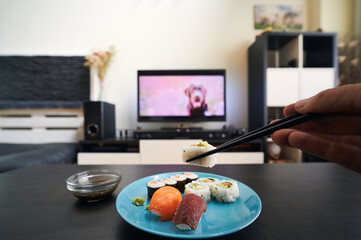 first person view of hand picking up a piece of sushi with chopsticks against an out-of-focus background of living room