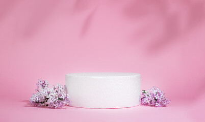 Cylindrical white podium on a pink background with hard shadows and lilac flowers. Minimal empty...