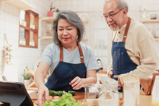 Portrait Of Mature Man And Woman Pensioner Using Internet Watching Teaching Cooking Video From The Tablet In The Kitchen At Home. Cheerful Asian Senior Couple Cooking Healthy Food Together