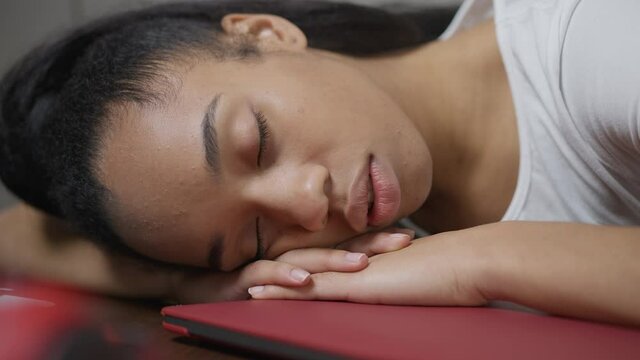 Close-up face of fatigued African American young woman sleeping on table in home office. Headshot of tired exhausted female freelancer napping indoors at work time. Multitasking and lifestyle