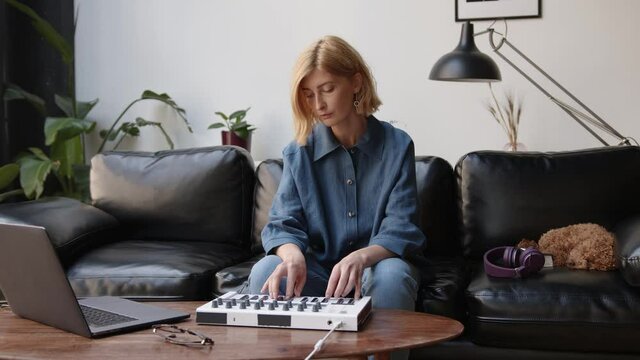 Young woman in casual clothes composing on midi controller, dog on the couch. Music producer using laptop. Student at online music school, songwriter