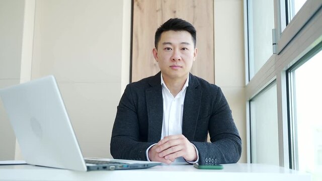 portrait of a confident asian business man who is seriously looking at the camera indoors. Office worker, employee male looks proud. Close up. Manager In workplace in formal suit and white shirt