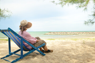 An elderly Asian woman in a hat sits on a deck chair by the beach. The concept of the elderly...