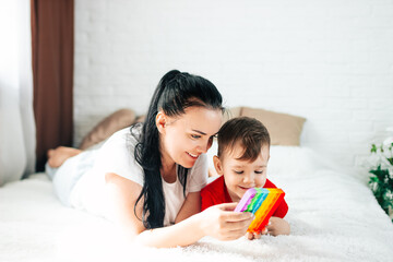 A mom and a 2-year-old boy play with a colorful fashion pop-it toy in a white room on the bed, an...
