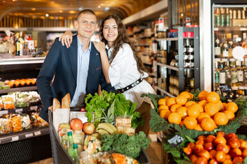 Portrait of young couple standing with full shopping cart while shopping in greengrocery