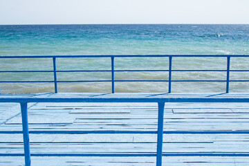 An old wharf covered with blue painted planks. The pier is fenced in front and rear with a blue, metal fence. In the background there is a blue sea and a bright sky. Copy space.