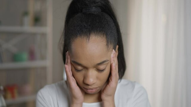 Close-up overburdened exhausted African American female freelancer rubbing temples standing in home office. Tired young woman having headache migraine looking at camera with sad facial expression
