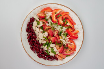 Boiled red beans, pieces of boiled chicken fillet, fresh chopped tomatoes, white onion and red sweet pepper, sprinkled with chopped parsley and dill in a round white plate on a white background.
