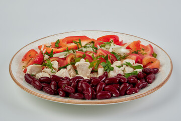 Boiled red beans, pieces of boiled chicken fillet, fresh chopped tomatoes, white onion and red sweet pepper, sprinkled with chopped parsley and dill in a round white plate on a white background.