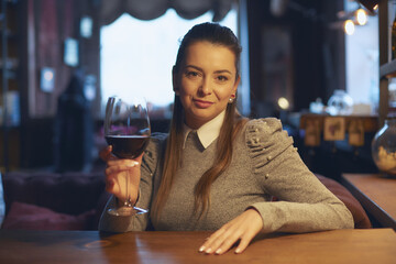 A beautiful young woman sits at a wooden table in the premises of a wine bar and drinks wine from a glass.