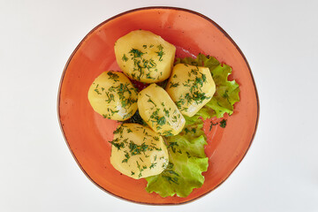 Boiled five pieces of potatoes, sprinkled with chopped dill with two leaves of green lettuce in a round orange plate on a white background.