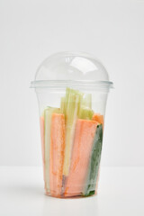 Transparent plastic cup with a lid with sliced strips of fresh carrots, cucumbers and celery on a white background.