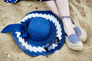 Women's feet in striped sandals, blue hat and sunglasses on the sand.