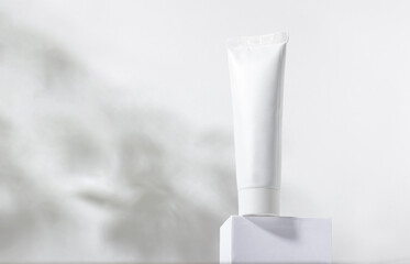 White cosmetic tube on a stand with leaves and shadows. Toothpaste, face and body cream. Female cosmetic tube with skin care product. Organic cosmetics. Copy space.