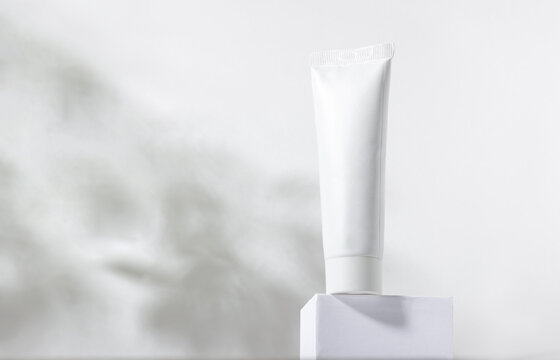 White cosmetic tube on a stand with leaves and shadows. Toothpaste, face and body cream. Female cosmetic tube with skin care product. Organic cosmetics. Copy space.