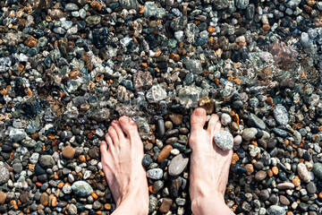 Female feet in sea water on a pebble shore top view in summer, many pebbles, copy space, relaxation in nature, swimming in the sea