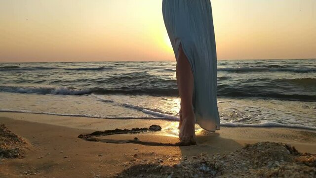 A young girl in blue long skirt is drawing a heart on sand of beach bootlessly. Sunrise.