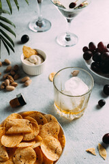 Whiskey is poured into a glass with ice. there are chips, glasses and other snacks nearby