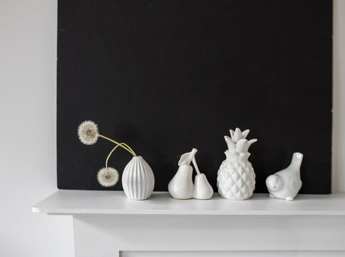 White figurines of pineapple, pear, bird, a vase with two dandelions on the background of a black painting on a white table.