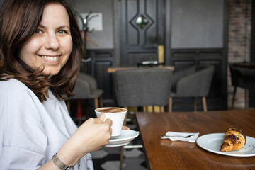 A woman sits at a table in a deserted cafe holding a cup of cappuccino and smiling. There is a plate with a croissant on an oak table