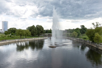 Fototapeta premium A large fountain with high jets of water in the middle of the river in a deserted city park under the cloudy sky in pandemic period