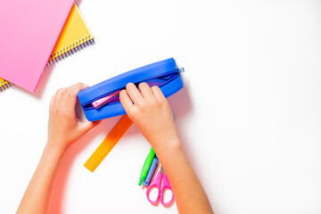 Child's hand keeping all the pencils, pens, scissors, etc. in his pencil case. Colorful notebooks on white table. Back to school concept