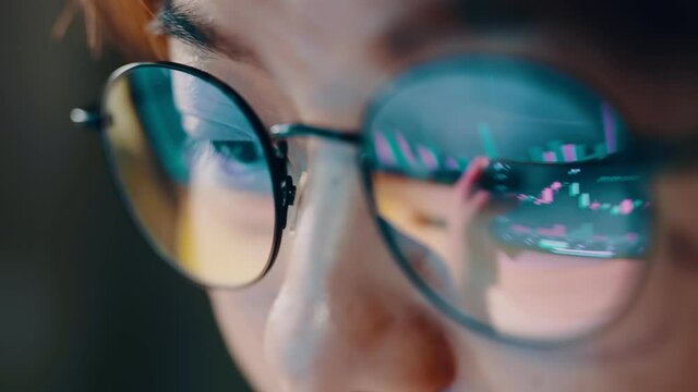 Closeup - Female eyes in eyeglasses late at night scrolling in front of laptop. Coder, she checking Bitcoin price chart on digital exchange on smartphone, cryptocurrency future price action prediction