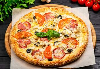Traditional Italian uncut whole pizza with chicken breast, ham, mushrooms, tomatoes, mozzarella cheese, olives, basil leaf. Pizza served on parchment paper on wooden board Dark background Food concept