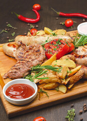 Grilled meat platter. pork steak, chicken steak, chicken sausages, peasant potatoes with garlic and herbs, grilled tomatoes, garlic sauce, BBQ sauce served on wooden board, ingredients on background
