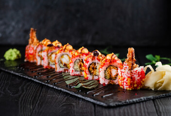 Japanese crunchy sushi roll on slate stone plate, wooden background. pieces with cucumber, tempura shrimp in rice with red flying fish roe on top served with sauce, ginger slices, wasabi and greens
