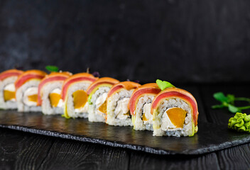 Side view of Japanese sushi roll on black slate stone plate on wooden background. sushi pieces with mango, cream cheese wrapped in rice with tuna, avocado on top served with sprouts of fresh herbs

