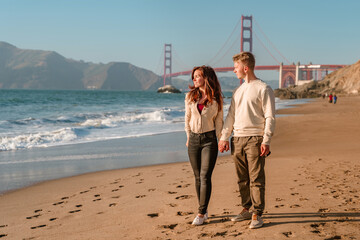 A young man and a woman take a romantic walk on the beach overlooking the Golden Gate Bridge at...