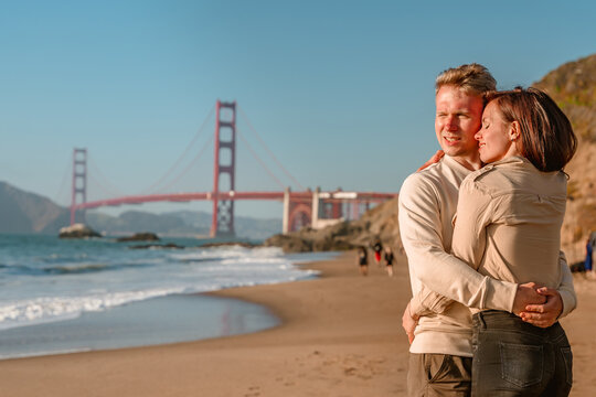 A young couple in love, a man and a woman, embrace on the beach in front of the Golden Gate Bridge in San Francisco