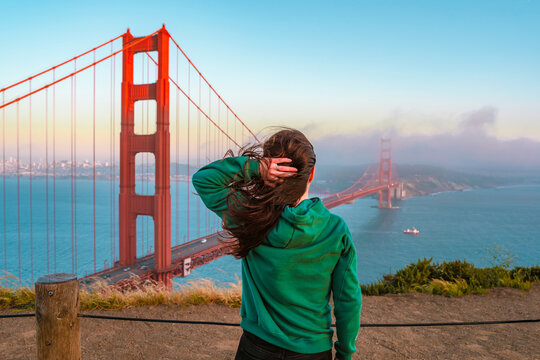 A young woman in a green hoodie stands on a hill overlooking the Golden Gate Bridge during sunset, San Francisco