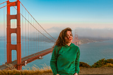 A young woman in a green hoodie stands on a hill overlooking the Golden Gate Bridge during sunset,...
