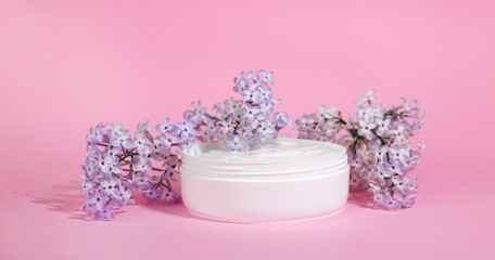 White jar of moisturizing face cream with pink cap isolated on pink background with lilac flowers. eco cosmetology or skin care concept.