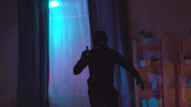 Funny thief in balaclava with a broken flashlight in the apartment
