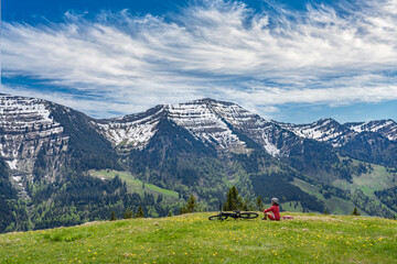 smiling senior woman riding her electric mountain bike on a sunny day in early spring with yello flowers on the meadows below the snow capped mountains of Nagelfluh chain near Oberstaufen, Allgaeu 