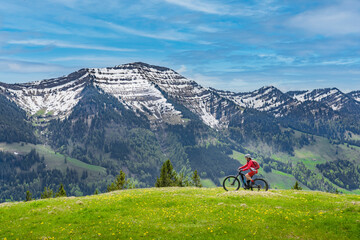 smiling senior woman riding her electric mountain bike on a sunny day in early spring with yello flowers on the meadows below the snow capped mountains of Nagelfluh chain near Oberstaufen, Allgaeu 