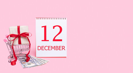 A gift box in a shopping trolley, dollars and a calendar with the date of 12 december on a pink background.