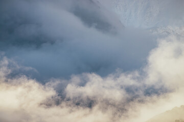 Minimalist mountain landscape with high snowy mountain wall in thick low clouds in golden sunshine. Awesome minimal scenery with dense low clouds in gold sunlight and great snow-covered mountains.