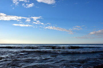 Obraz premium Waves on the evening sea before sunset under a blue sky with light clouds, which themselves begin to cast light shadows on the sky.